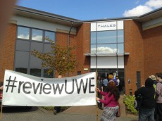 People holding a '#reviewUWE' banner outside Thales offices