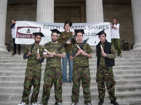Students in camouflage and mortar boards hold graduation certificates and fake guns in front of a 'ditch the arms shares' banner