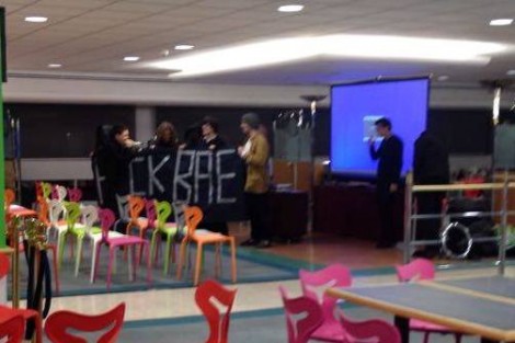 Students hold an anti-BAE banner in a room with empty chairs laid out in rows. Two people pack up presentation equipment.