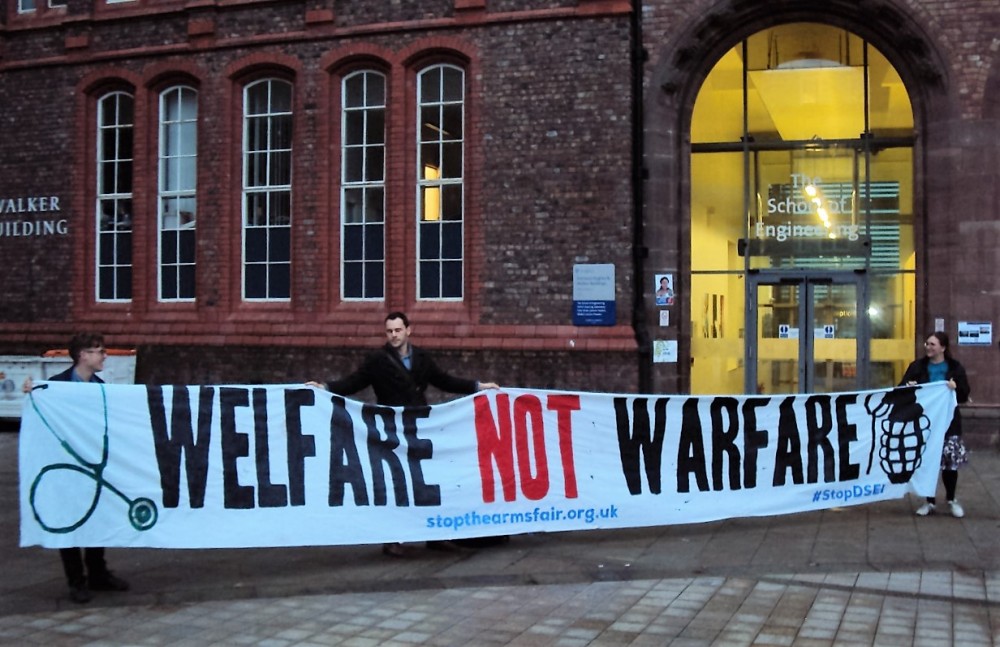 Three students hold a banner reading "Welfare not Warfare" in front of a building signed as "The School of Engineering".